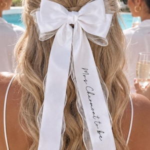 May include: A white hair bow made of satin and sheer organza fabric, featuring two long trailing ribbons. One ribbon is printed with the black cursive text "Mrs Charmont to be". The bow is secured at the centre with a matching satin knot.