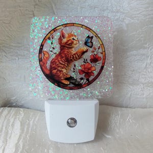 May include: A decorative night light with a square, glitter-covered panel. It features a circular image of an orange kitten reaching for a blue butterfly, surrounded by red flowers. The base is white.