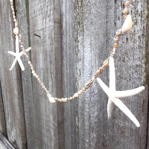 May include: A decorative garland featuring white starfish and small seashells strung together. The garland is suspended against a weathered wooden background, creating a coastal or nautical theme. The starfish are approximately 4 inches in diameter.