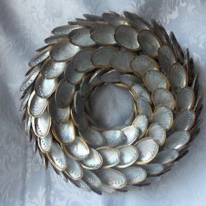 May include: A circular wreath composed of iridescent, oval shells, arranged in a spiral design. The shells display a pearlescent sheen with touches of blue, green, and gold. The wreath is set against a white backdrop.