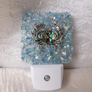 Könnte beinhalten: Ein blaues und weißes Nachtlicht mit einer silbernen Krabbe mit einem schillernden Panzer. Das Nachtlicht besteht aus zerbrochenem Glas und Glitzer.