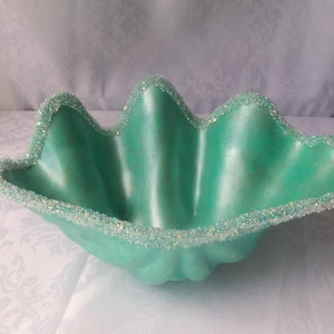 Puede incluir: Un cuenco decorativo verde mar con borde festoneado. El borde está incrustado con purpurina iridiscente. El cuenco tiene un acabado liso y brillante, probablemente hecho de cerámica o material similar.