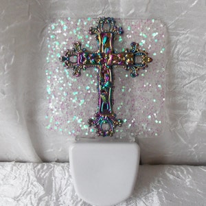 May include: A white night light with an iridescent cross on a glittery background. The cross has a floral design and is made of metal.