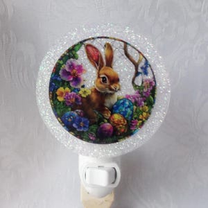 May include: A decorative night light featuring a circular design with a brown bunny surrounded by colorful flowers and Easter eggs. The night light has a white base and a glittery border.