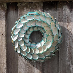 Peut inclure: Une couronne décorative composée de coquillages irisés bordés de turquoise. Les coquillages sont disposés en cercle, créant un design unique. La couronne est fixée sur une surface en bois.