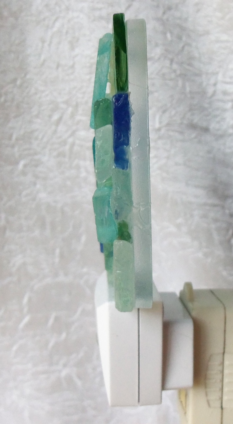 K&ouml;nnte beinhalten: Ein Nachtlicht mit einem Mosaik aus blauen, gr&uuml;nen und wei&szlig;en Glasst&uuml;cken. Das Licht ist an einem wei&szlig;en Stecker befestigt.