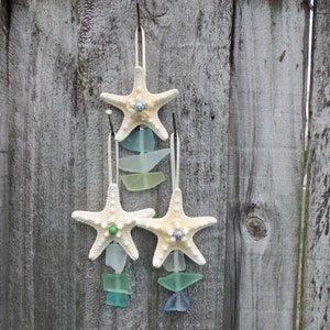 May include: Three white starfish ornaments with blue and green glass beads hanging from a wooden fence.