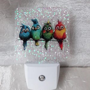 Puede incluir: Luz de noche decorativa con cuatro pájaros de dibujos animados coloridos posados en una rama. Los pájaros son azules, verdes y rojos, sobre un fondo brillante. La base es blanca con una pequeña luz redonda.