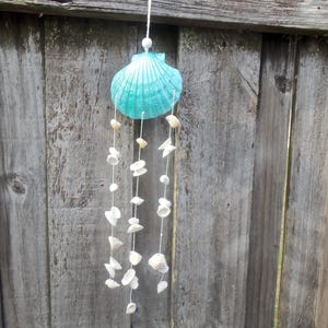 May include: A turquoise seashell wind chime with strands of small white seashells hanging from it. The wind chime is suspended from a white string against a weathered wooden fence.