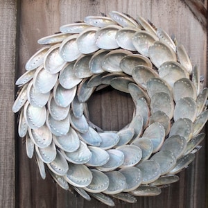 May include: A circular wreath made of iridescent, pearlescent seashells. The shells are arranged in overlapping layers, creating a textured, decorative piece. The wreath is displayed against a weathered wooden background, adding a rustic touch.