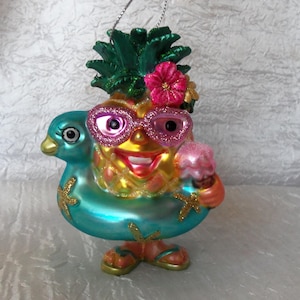 May include: A glass ornament shaped like a pineapple wearing sunglasses and a blue inflatable ring, holding an ice cream cone. The pineapple has a pink flower on its head and is decorated with glitter.
