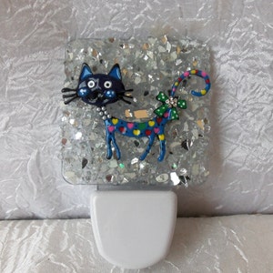 Op de afbeelding: Een zilveren glitter nachtlampje met een blauwe kat met een kleurrijk hartpatroon en een strik op zijn staart.