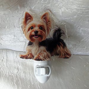 Op de afbeelding: Een decoratief nachtlampje met een Yorkshire Terrier hond. De hond heeft bruin, zwart en bruin haar, met een vrolijke uitdrukking. Het nachtlampje is wit en heeft een aan/uit-schakelaar.