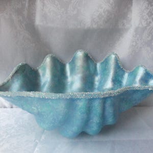 May include: A decorative, light blue clam shell-shaped bowl with a glittery, silver-colored rim. The bowl has a textured surface and a wavy edge. The interior has a pearlescent sheen. A unique accent piece for home decor.