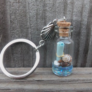 Puede incluir: Un llavero de plata con un pequeño encanto de botella de vidrio. La botella contiene un trozo de papel enrollado, conchas marinas y pequeñas piedras. La botella está coronada con un corcho y un encanto de concha de mar de plata.