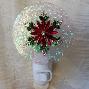 May include: A white night light with a clear, round, glittery cover featuring a red poinsettia with green leaves and rhinestones.