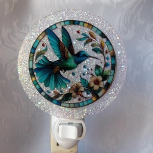 Puede incluir: Luz de noche decorativa con un diseño de colibrí. El colibrí está representado con plumas azules y verdes, rodeado de flores y hojas. La luz de noche tiene un borde redondo brillante y un interruptor de encendido/apagado blanco.