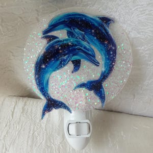 May include: A night light featuring two blue dolphins with a starry design, set against a glittery background. The dolphins are in a swimming pose. The night light has a white base with an on/off switch.