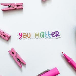 Op de afbeelding: Een witte sticker met de tekst "you matter" in regenboogkleuren. De sticker bevindt zich op een witte achtergrond met roze wasknijpers, een roze hart, een roze pen en een rode stift.