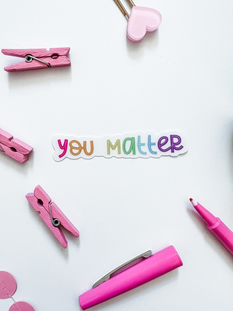 Op de afbeelding: Een witte sticker met de tekst "you matter" in regenboogkleuren. De sticker bevindt zich op een witte achtergrond met roze wasknijpers, een roze hart, een roze pen en een rode stift.