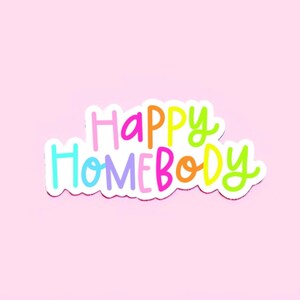 May include: A white sticker with the text "Happy Homebody" in rainbow colors. The sticker has a white outline and is on a pink background.