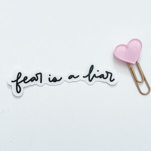 May include: A white sticker with the text "fear is a liar" in black cursive writing. A pink heart-shaped paperclip is attached to the sticker.
