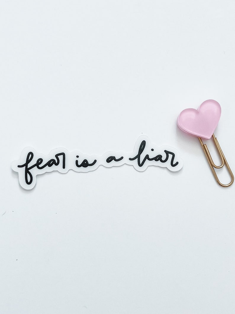 May include: A white sticker with the text "fear is a liar" in black cursive writing. A pink heart-shaped paperclip is attached to the sticker.