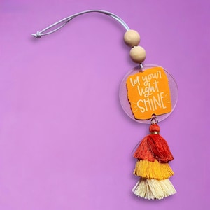 May include: A decorative car charm with a clear acrylic disc and an orange insert that reads "Let your light SHINE". The charm has a layered tassel in red, yellow, and cream colors, and wooden beads. The charm hangs from a silver cord.