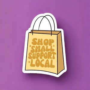 May include: A white sticker with a brown paper bag illustration. The bag has a black handle and the text "SHOP SMALL SUPPORT LOCAL" in yellow.