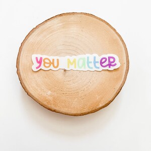 Op de afbeelding: Een regenboogkleurige sticker met de tekst "you matter" op een houten schijf.