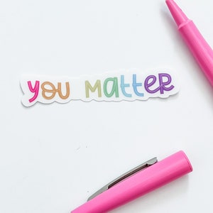 Op de afbeelding: Een witte sticker met de woorden "you matter" geschreven in regenboogkleuren. De sticker is op een witte achtergrond met een roze markeerstift.