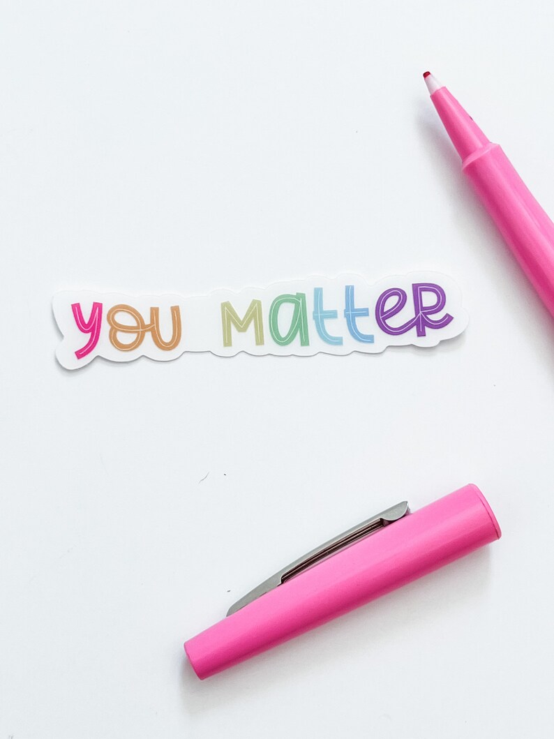 Op de afbeelding: Een witte sticker met de woorden "you matter" geschreven in regenboogkleuren. De sticker is op een witte achtergrond met een roze markeerstift.