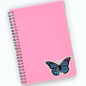 May include: A pink spiral bound notebook with a blue butterfly sticker on the cover. The butterfly sticker has the text "This is my everything" on it.