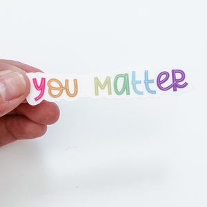 Op de afbeelding: Een sticker met de tekst "you matter" in regenboogkleuren. De sticker is op een witte achtergrond.