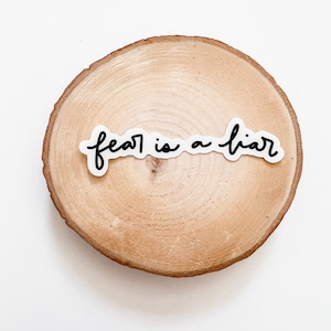 May include: A white sticker with black lettering on a wooden round slice. The sticker reads "fear is a liar".