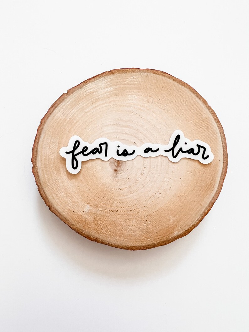 May include: A white sticker with black lettering on a wooden round slice. The sticker reads "fear is a liar".