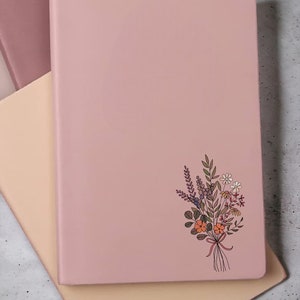 May include: A pink notebook with a floral design on the front cover. The notebook is closed and sitting on a light brown surface.