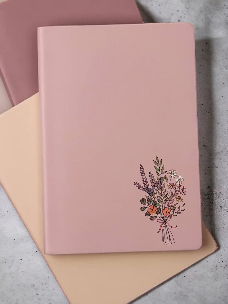 May include: A pink notebook with a floral design on the front cover. The notebook is closed and sitting on a light brown surface.