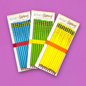 May include: Three packs of pencils in blue, green, and yellow, each held together with a colored band. The pencils are on white cardstock with the text "Positively Lettering" and "www.positivelylettering.com". Each pencil has text printed on it.