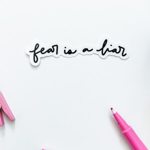 May include: A white sticker with the text "fear is a liar" written in black cursive script. The sticker is on a white background with pink clothespins and pink markers.