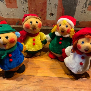 Puede incluir: Cuatro juguetes de peluche vestidos con ropa de invierno. Los juguetes sonríen y tienen las mejillas sonrosadas. Están de pie sobre un suelo de madera.
