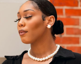 May include: A pearl necklace and matching stud earrings. The necklace features round, white pearls, and the earrings are large, round pearls. The jewellery is worn with a black top, set against a brick wall background.