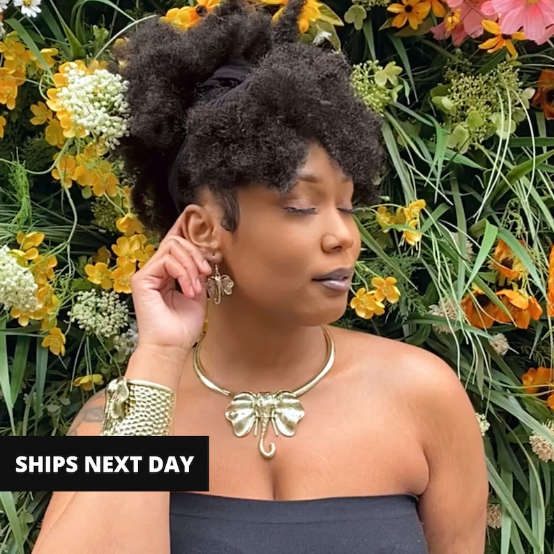 May include: A woman with dark curly hair is wearing a gold elephant pendant necklace and matching earrings. The necklace is a choker style and the pendant is large and detailed. The woman is wearing a black strapless top and is standing in front of a wall of flowers.