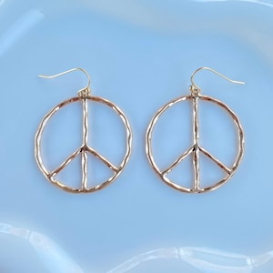 May include: A pair of gold-colored peace sign earrings. Each earring features a hammered texture around the circle and the peace symbol's vertical and diagonal lines. The earrings have a hook closure.