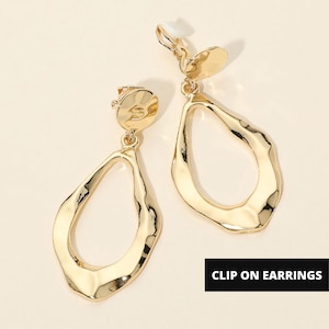 May include: A pair of gold-toned clip-on earrings featuring a textured, irregular teardrop-shaped pendant suspended from a circular stud. The metal surface has a polished, reflective finish with organic, wavy edges. The text CLIP ON EARRINGS is visible.
