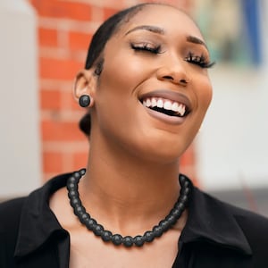 May include: A black beaded necklace and matching stud earrings. The necklace is made of round, dark beads. The earrings are simple, round studs. The jewelry is worn with a black top.