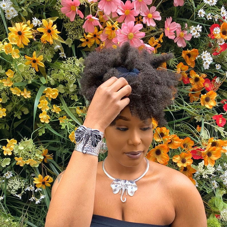 May include: A silver elephant-themed jewelry set, including a cuff bracelet and a necklace. The cuff bracelet features a black and silver elephant design. The necklace has a matching elephant pendant. The background is a vibrant display of colorful flowers.