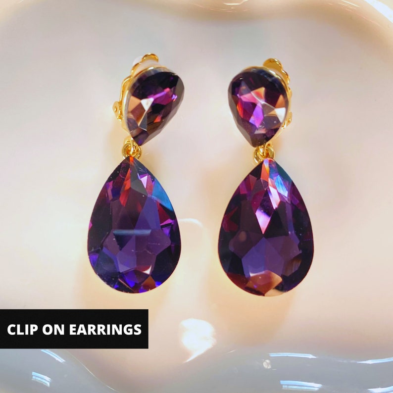 May include: A pair of clip-on earrings with large teardrop-shaped purple crystals. The earrings have a gold-tone finish.