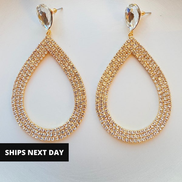 May include: A pair of gold-toned, teardrop-shaped earrings encrusted with small, sparkling crystals. Each earring has a large, clear, teardrop-shaped crystal at the top. The earrings are displayed on a white surface.