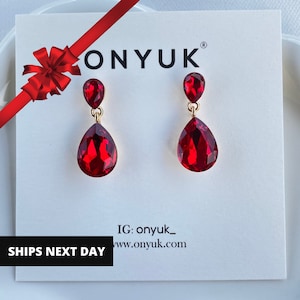 May include: A pair of red teardrop earrings with gold-colored accents. The earrings are displayed on a white card with the brand name "ONYUK" and the text "SHIPS NEXT DAY". A red ribbon and bow are in the upper left corner.